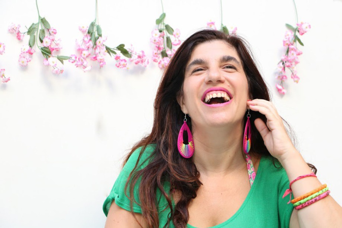 Luciana Peker. Fuente: Wikipedia Luciana Peker se retrata en una foto. Es una mujer de cabello cafe obscuro, y largo, más abajo de sus hombros.  Está sonriendo con vista hacía al horizonte y arriba, mano levantada a la mejilla de su cara, con sonrisa grande y boca abierta. Viste una camiseta verde con aretes grandes y pulseras de muchos colores. En el fondo contra pared hay seis ramitas de flores color rosa pálida con hojas verdes, colgadas cada rama de manera deocrativa, hojas y pétalos hacía abajo.