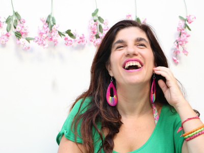 Luciana Peker. Fuente: Wikipedia Luciana Peker se retrata en una foto. Es una mujer de cabello cafe obscuro, y largo, más abajo de sus hombros.  Está sonriendo con vista hacía al horizonte y arriba, mano levantada a la mejilla de su cara, con sonrisa grande y boca abierta. Viste una camiseta verde con aretes grandes y pulseras de muchos colores. En el fondo contra pared hay seis ramitas de flores color rosa pálida con hojas verdes, colgadas cada rama de manera deocrativa, hojas y pétalos hacía abajo.